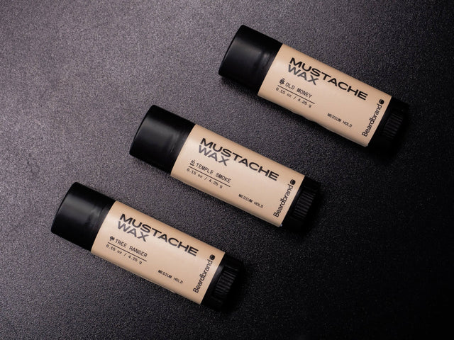 Three Beardbrand Mustache Waxes laid on a textured black surface in a diagonal line in fragrances Old Money, Temple Smoke and Tree Ranger.