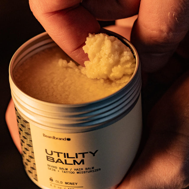 A person holds an open container of Beardbrand Utility Balm while scooping the product out with one finger.