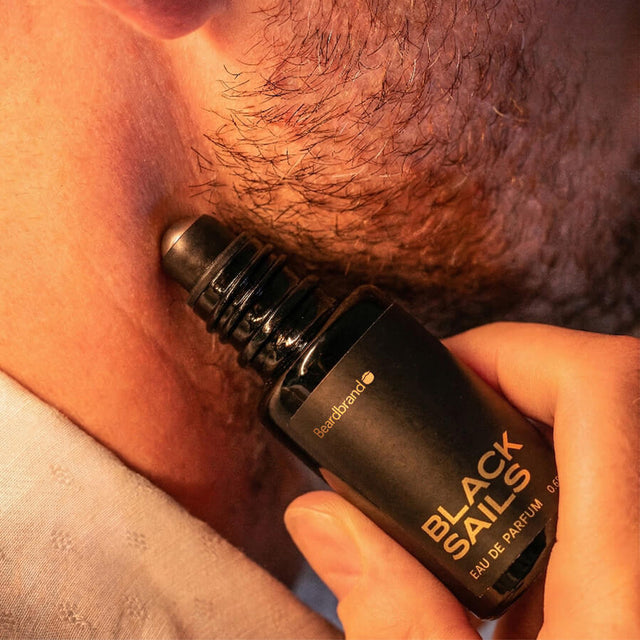Close-up of stubbled lower face with Beardbrand Black Sails Eau de Parfum roll-on bottle held against the skin.