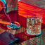 Glass of whiskey with ice on a wooden table, accompanied by a smoking cigar in an ashtray. A wooden box sits in the background, a leather wallet lies in the foreground.