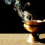 Golden brass incense burner with wisps of white and blue smoke rising against a black background. The ornamental vessel sits on a light beige surface, casting a soft shadow.