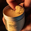 A person holds an open container of Beardbrand Beard Balm while scooping the product out with one finger.